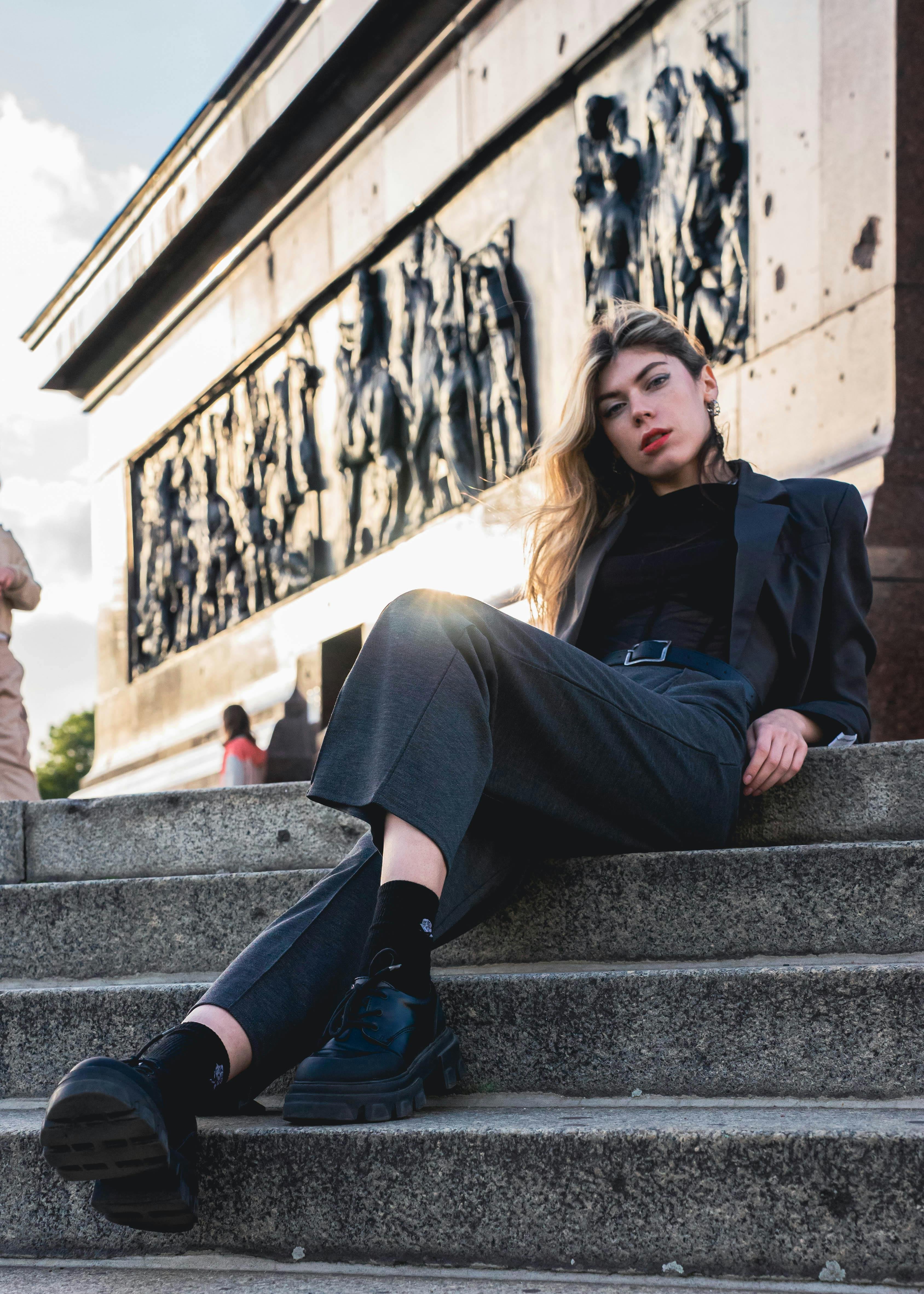 Woman sitting on steps in an urban setting with a large architectural structure in the background

